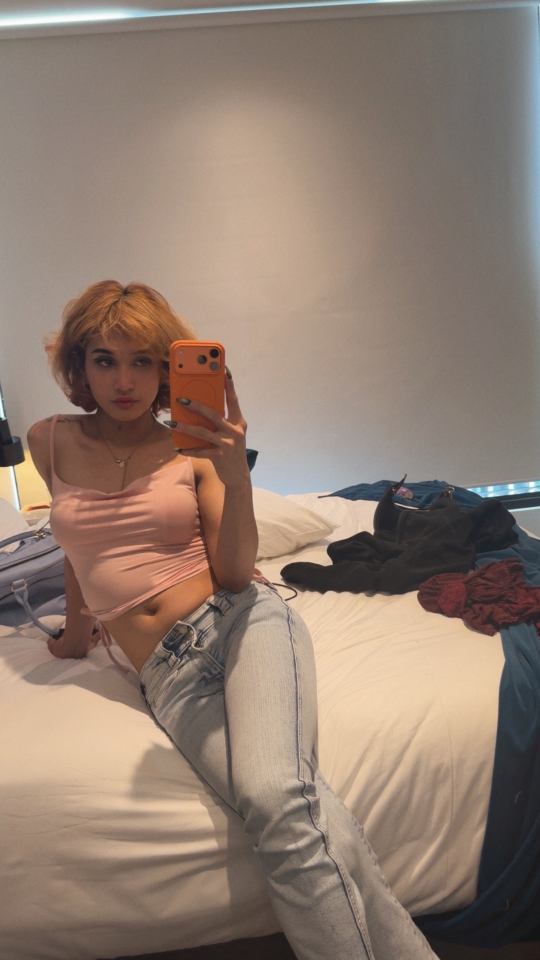 POR$CHA in pink top and jeans seated on a bed, mirror self-portrait