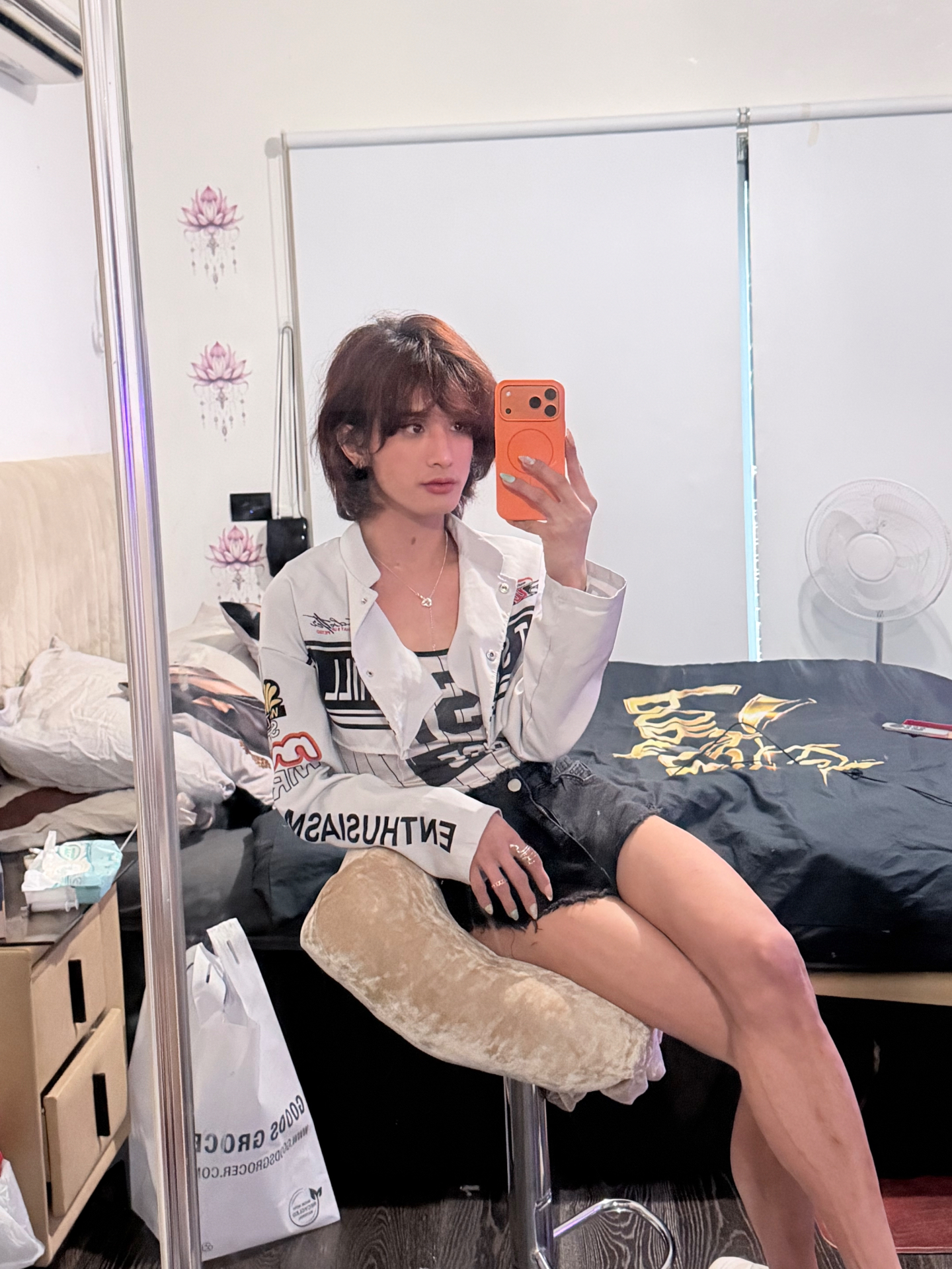 POR$CHA seated in a white graphic jacket and dark shorts, mirror self-portrait