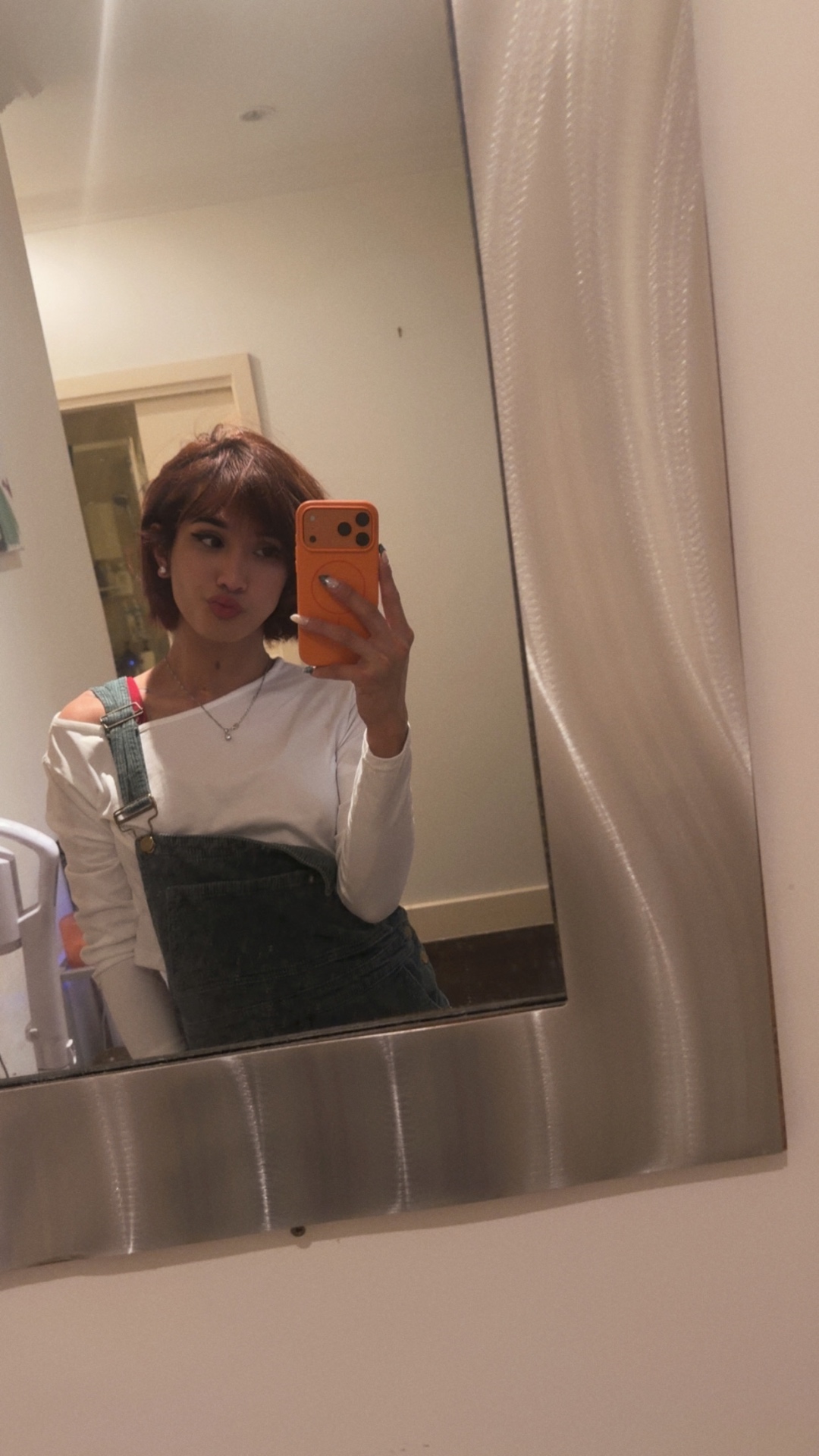 POR$CHA in white top and denim overalls, bathroom mirror self-portrait